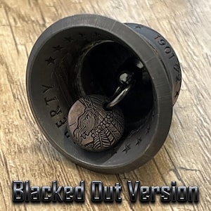 May include: A dark-toned metal bell featuring a circular clapper with an embossed profile design. The interior rim of the bell is engraved with the word LIBERTY, a series of stars, and the year 1901. The exterior surface has a matte black finish.