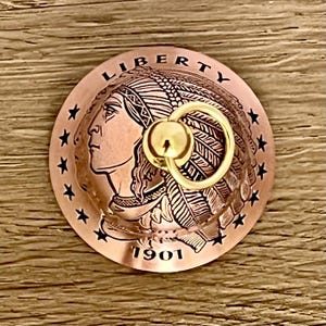 May include: A circular copper-toned metal object featuring a relief profile of a person wearing a feathered headdress, the word LIBERTY, the year 1901, and a ring of stars. A small, polished gold-colored metal ring is attached to the center.