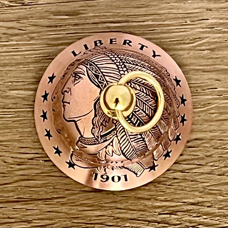May include: A circular copper-toned metal object featuring a relief profile of a person wearing a feathered headdress, the word LIBERTY, the year 1901, and a ring of stars. A small, polished gold-colored metal ring is attached to the center.
