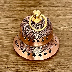 May include: A copper-colored bell featuring an engraved design of feathers and stars. The bell has a shiny gold-toned ring at the top and includes the text 1901 and LIBERTY etched into the surface. The design wraps around the bell's exterior.