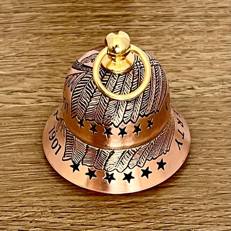 May include: A copper-colored bell featuring an engraved design of feathers and stars. The bell has a shiny gold-toned ring at the top and includes the text 1901 and LIBERTY etched into the surface. The design wraps around the bell's exterior.