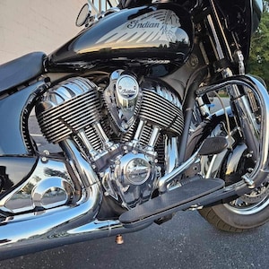 May include: A black Indian motorcycle featuring a prominent V-twin engine with polished chrome cooling fins and exhaust pipes. The fuel tank displays the Indian brand logo and a graphic of a Native American profile. Chrome engine covers show the Indian name.
