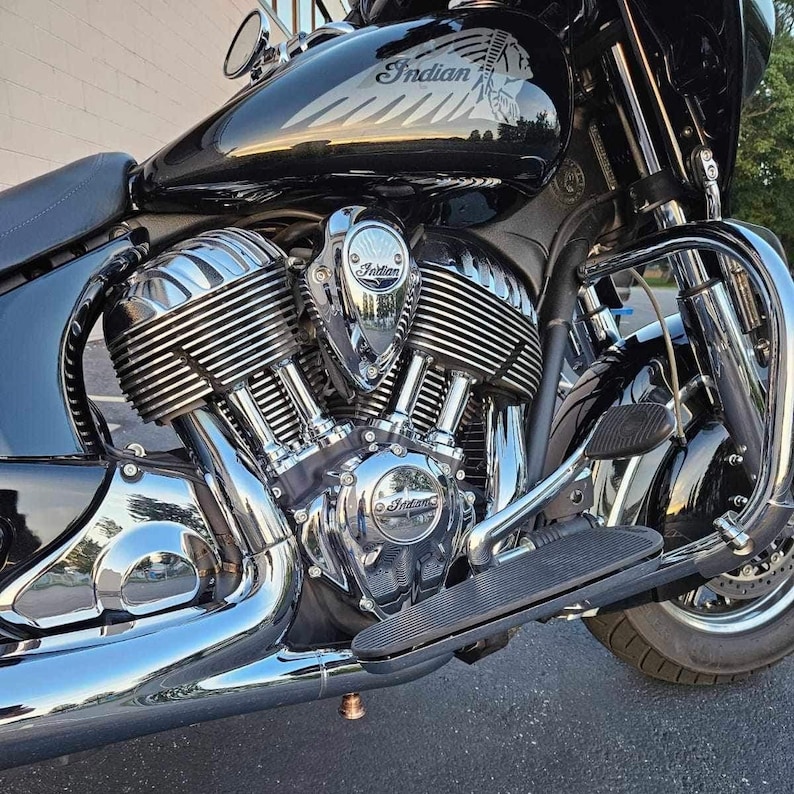 May include: A black Indian motorcycle featuring a prominent V-twin engine with polished chrome cooling fins and exhaust pipes. The fuel tank displays the Indian brand logo and a graphic of a Native American profile. Chrome engine covers show the Indian name.