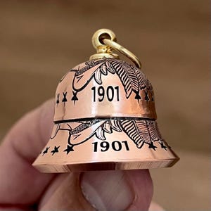 May include: A copper-colored bell charm featuring a gold-toned top loop. The bell is engraved with black leaf patterns, small star motifs, and the number 1901 repeated twice on the surface. The design is symmetrical with a flared bottom edge.