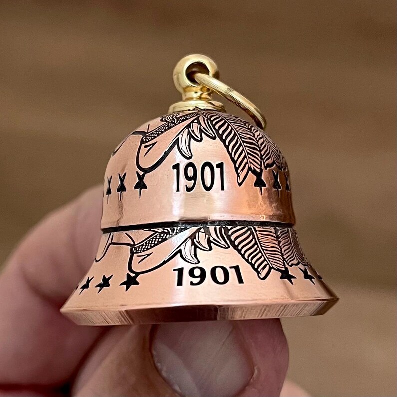 May include: A copper-colored bell charm featuring a gold-toned top loop. The bell is engraved with black leaf patterns, small star motifs, and the number 1901 repeated twice on the surface. The design is symmetrical with a flared bottom edge.