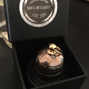 May include: A small, black box with a gold skull-shaped bell inside. The box has a label that reads "Coin and Spoon Rings Cents of Beauty Est. 2014".