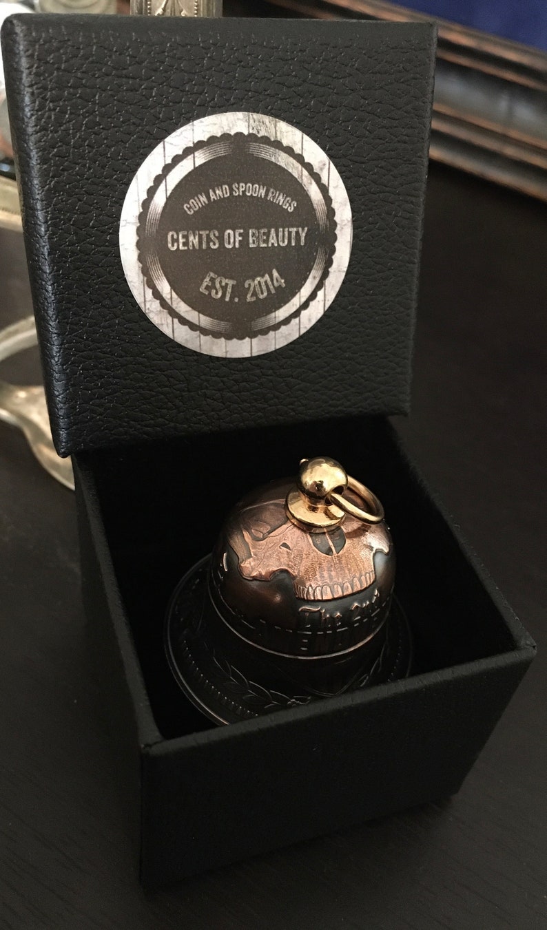 May include: A small, black box with a gold skull-shaped bell inside. The box has a label that reads "Coin and Spoon Rings Cents of Beauty Est. 2014".