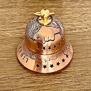 May include: A copper-colored bell featuring an embossed profile of a person wearing a feathered headdress, reminiscent of a coin design. The word LIBERTY and the year 1907 are engraved on the side, accented by a row of cut-out stars and a gold-toned handle.