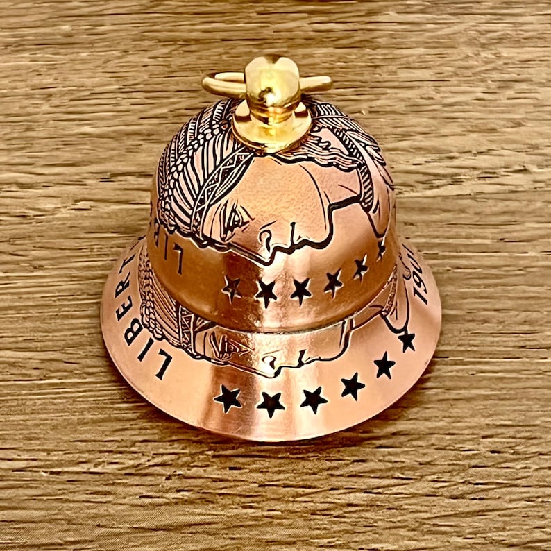 May include: A copper-colored bell featuring an embossed profile of a person wearing a feathered headdress, reminiscent of a coin design. The word LIBERTY and the year 1907 are engraved on the side, accented by a row of cut-out stars and a gold-toned handle.