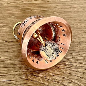 May include: A copper-toned bell crafted from a coin, featuring the year 1901 and the word LIBERTY embossed on the exterior. The bell includes a clapper made from a smaller coin depicting a profile of a person wearing a feathered headdress.