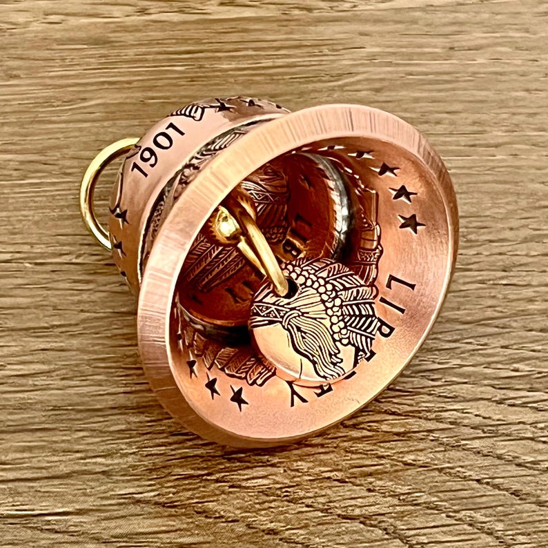 May include: A copper-toned bell crafted from a coin, featuring the year 1901 and the word LIBERTY embossed on the exterior. The bell includes a clapper made from a smaller coin depicting a profile of a person wearing a feathered headdress.