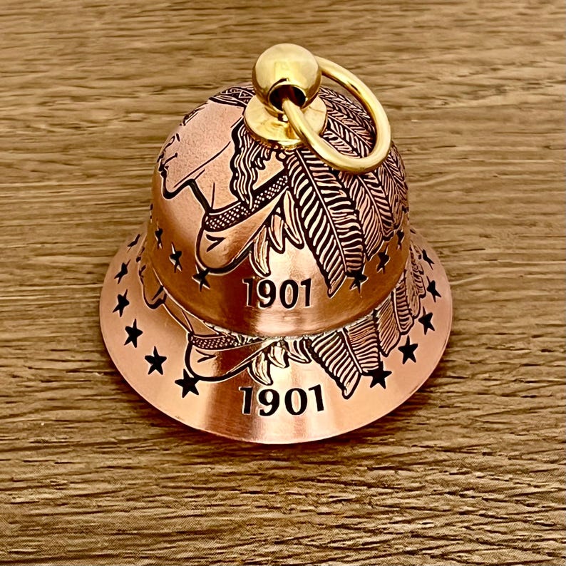 May include: A copper-colored bell featuring an engraved profile of a person wearing a feathered headdress, with the year 1901 printed twice and a series of black stars encircling the base. A gold-toned metal ring is attached to the top.