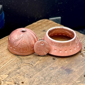 May include: A copper-colored metal dome, a small circular disc, and a ring, all featuring embossed details of a profile with a feathered headdress and stars. The dome is stamped with the year 1901 and has a small hole at the top center.