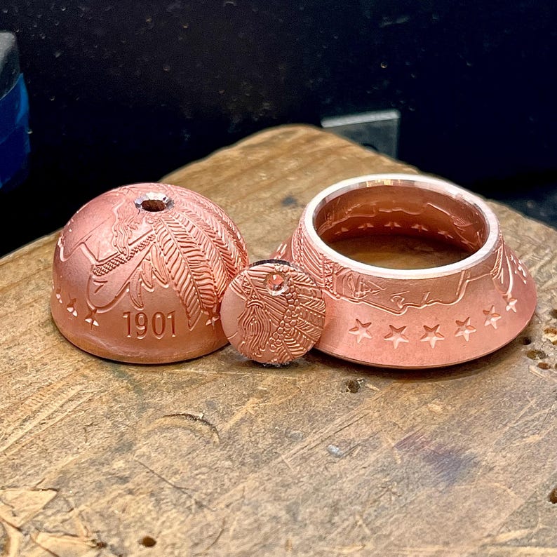 May include: A copper-colored metal dome, a small circular disc, and a ring, all featuring embossed details of a profile with a feathered headdress and stars. The dome is stamped with the year 1901 and has a small hole at the top center.