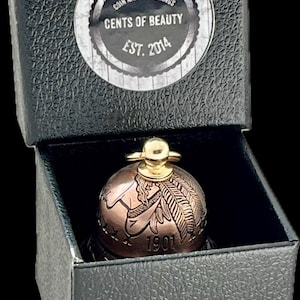 May include: A copper-toned bell crafted from a 1901 coin, featuring a gold-colored top handle, housed in a black textured box. The box lid displays a circular label reading COIN AND SPOON RINGS CENTS OF BEAUTY EST. 2014 in white and black text.