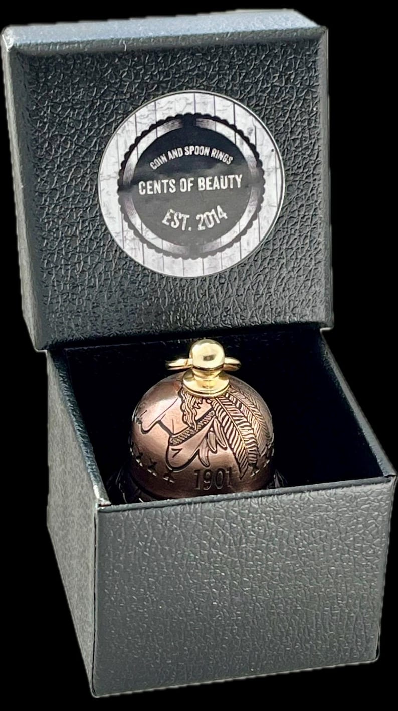 May include: A copper-toned bell crafted from a 1901 coin, featuring a gold-colored top handle, housed in a black textured box. The box lid displays a circular label reading COIN AND SPOON RINGS CENTS OF BEAUTY EST. 2014 in white and black text.