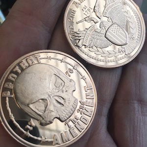 May include: Two copper-colored challenge coins. The first coin features a skull with barbed wire around the edge and the text "Protect the 2nd Amendment". The second coin features an eagle with a shield and the text "The 2nd Amendment est 1791 Shall Not Be Infringed".