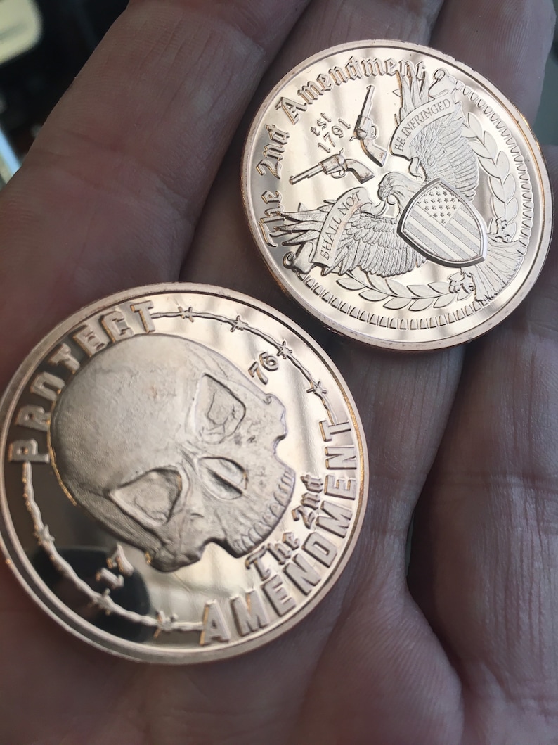 May include: Two copper-colored challenge coins. The first coin features a skull with barbed wire around the edge and the text "Protect the 2nd Amendment". The second coin features an eagle with a shield and the text "The 2nd Amendment est 1791 Shall Not Be Infringed".