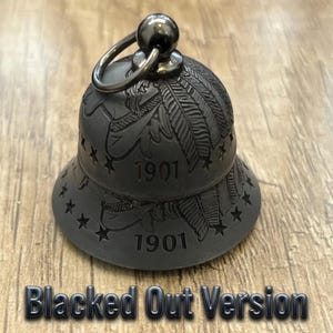May include: A matte black metal bell featuring an engraved feather design, a ring of small stars around the base, and the number 1901 etched into the surface. A circular metal loop is attached to the top of the bell for hanging purposes.