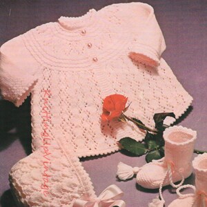 May include: A light pink knitted baby set, including a sweater with pearl buttons, a bonnet, and booties with tassels. A red rose and a pink ribbon are also in the image. The text "KnittingUpVintage" is on the bonnet.