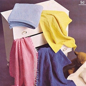 Könnte beinhalten: Vier gestrickte Babydecken in verschiedenen Farben: hellblau, gelb, rosa und lila. Die Decken sind gefaltet und auf einer weißen Kommode ausgestellt.
