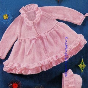Könnte beinhalten: Ein rosa gestricktes Baby-Jäckchen und eine passende Mütze mit Rüschenbesatz und einer Schleife aus Satinband. Das Set ist aus zartem Garn gefertigt und das Jäckchen hat einen Knopfverschluss.