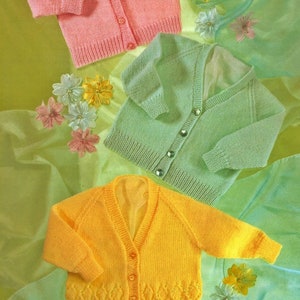 Puede incluir: Tres cárdigans de punto para bebé en rosa, verde y amarillo. Los cárdigans están dispuestos sobre un fondo de tela verde y blanca. El texto "EASY KNIT DESIGN" es visible en la esquina superior derecha de la imagen.