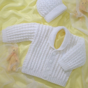 May include: A white knitted baby cardigan with a button fastening and a matching white knitted hat with a pom-pom on top.