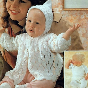 May include: A baby in a white knitted outfit, comprising a hat, cardigan, and trousers. The cardigan features a textured pattern with bobbles and diamond shapes. The baby is seated and held by an adult.