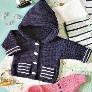 Könnte beinhalten: Marineblaue Babyjacke aus Strick mit Kapuze, weißen Streifen an Ärmeln und Taschen sowie weißen Knöpfen. Die Jacke ist Teil einer Kollektion gestrickter Babykleidung. Ein Spielzeug-Segelboot und fischförmige Dekorationen befinden sich im Hintergrund.
