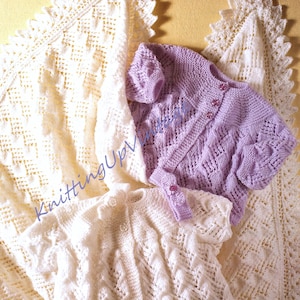 May include: Two knitted baby cardigans, one white and one lavender, with a white crocheted blanket in the background.