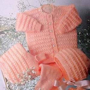 May include: A knitted baby outfit in a soft pink color. The set includes a cardigan, a hat, and booties. The cardigan has a button closure and a delicate stitch pattern.