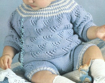 May include: A baby wearing a light blue knitted sweater and matching pants with a white trim. The sweater has a diamond pattern and the pants have a white trim around the legs. The baby is sitting on a white and blue blanket with a white lace trim.