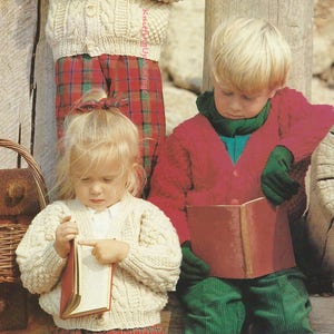 May include: Three children wearing knitted cardigans. The child on the left is wearing a cream cardigan with a cable knit pattern and holding binoculars. The child in the middle is wearing a cream cardigan with a cable knit pattern and reading a book. The child on the right is wearing a red cardigan and reading a book. 51-76 cm 20-30 ins