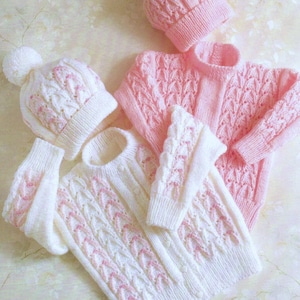 May include: Two knitted baby sweaters with pink and white cable knit patterns. One sweater is pink and the other is white with pink accents. Both sweaters have matching hats.