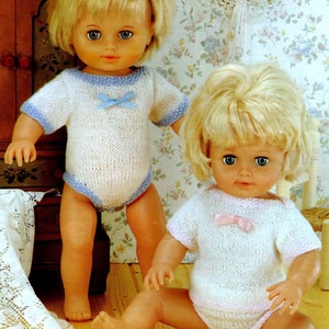 May include: Two blonde dolls wearing knitted outfits. The doll on the left is wearing a white knitted bodysuit with blue trim and a blue bow. The doll on the right is wearing a white knitted bodysuit with pink trim and a pink bow.