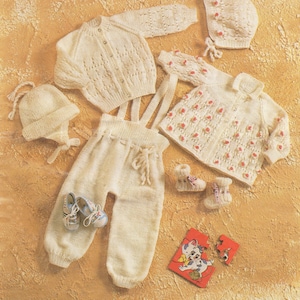 May include: A set of knitted baby clothes in white, including a cardigan, a hat, a pair of pants, a jacket, and booties. The jacket has pink flower accents.