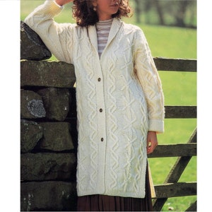 May include: A long, white knitted cardigan with a cable knit pattern and button fastening. The cardigan has a collar and is being worn by a person standing in front of a stone wall.