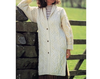 Könnte beinhalten: Ein langer, weißer Strickcardigan mit einem Zopfmuster und Knöpfen. Der Cardigan hat einen Kragen und wird von einer Person getragen, die vor einer Steinmauer steht.