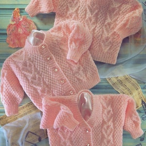 May include: Three pink knitted cardigans with button closures and cable knit patterns. The cardigans are arranged on a blue and white patterned surface.