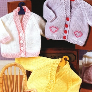 May include: Three knitted baby cardigans in pink, white and yellow with button fastenings. The cardigans are hanging on hangers in a wooden cabinet.