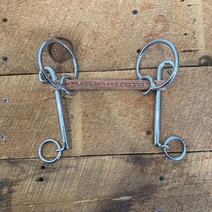 May include: A silver horse bit with a leather mouthpiece. The bit has two rings on each side and a centre bar.