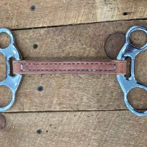 May include: A silver horse bit with a brown leather strap. The bit has two rings on each side.