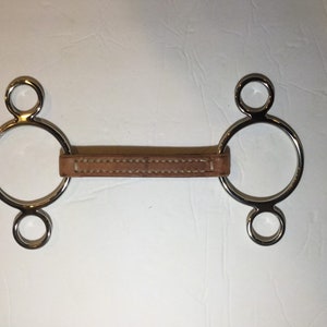 Puede incluir: Un bocado de caballo de plata con una correa de cuero. El bocado tiene tres anillos a cada lado.