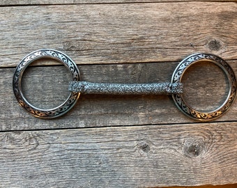 May include: A silver horse bit with decorative floral patterns on the rings and a grey braided centre. The bit is designed for equestrian use and is displayed on a weathered wooden surface.