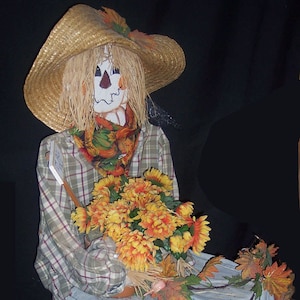 May include: A handmade scarecrow doll wearing a straw hat, a plaid shirt, and denim trousers. The scarecrow holds a bouquet of orange and yellow flowers and is decorated with autumn leaves. The doll has a painted face and a colourful scarf. A charming autumn decoration.