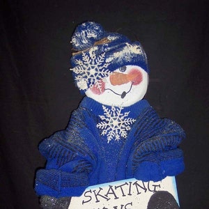 Op de afbeelding: Een grillige sneeuwpopfiguur gemaakt van blauwe stof, met een bijpassende hoed en trui. De sneeuwpop houdt een bord vast met de tekst "SKATING LESSONS". Witte sneeuwvlokaccenten en een wortelneus maken de winterse decoratie compleet.