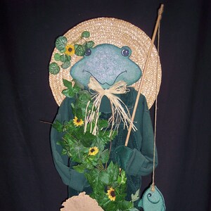 May include: A whimsical frog figurine wearing a straw hat, overalls, and holding a fishing rod with a fish. The frog is green with a straw hat and is decorated with sunflowers and vines. The base has the text: https://etsy.com/shop/happyhollowcreation.