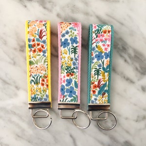 May include: Three keychains with floral fabric straps and silver key rings. The straps are yellow, pink, and teal with a white background and colorful floral patterns.