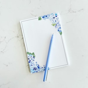 May include: A white notepad with a black border and blue floral designs on the corners. A light blue pen is lying on top of the notepad.